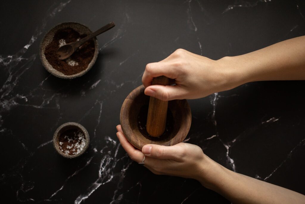 From above of crop unrecognizable female using pestle while grinding fresh aromatic coffee in mortar on black marble table