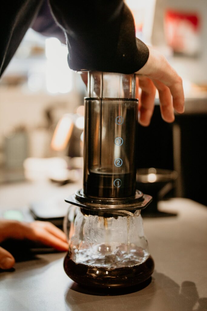 Unrecognizable skilled barista pressing professional aeropress while preparing aromatic coffee into glass pot while working in cafe on blurred background