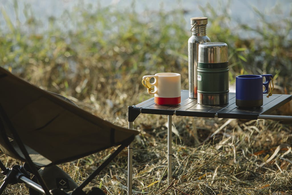 Stainless steel vacuum flasks and mugs with hot drinks on portable table near folding chair on grass shore