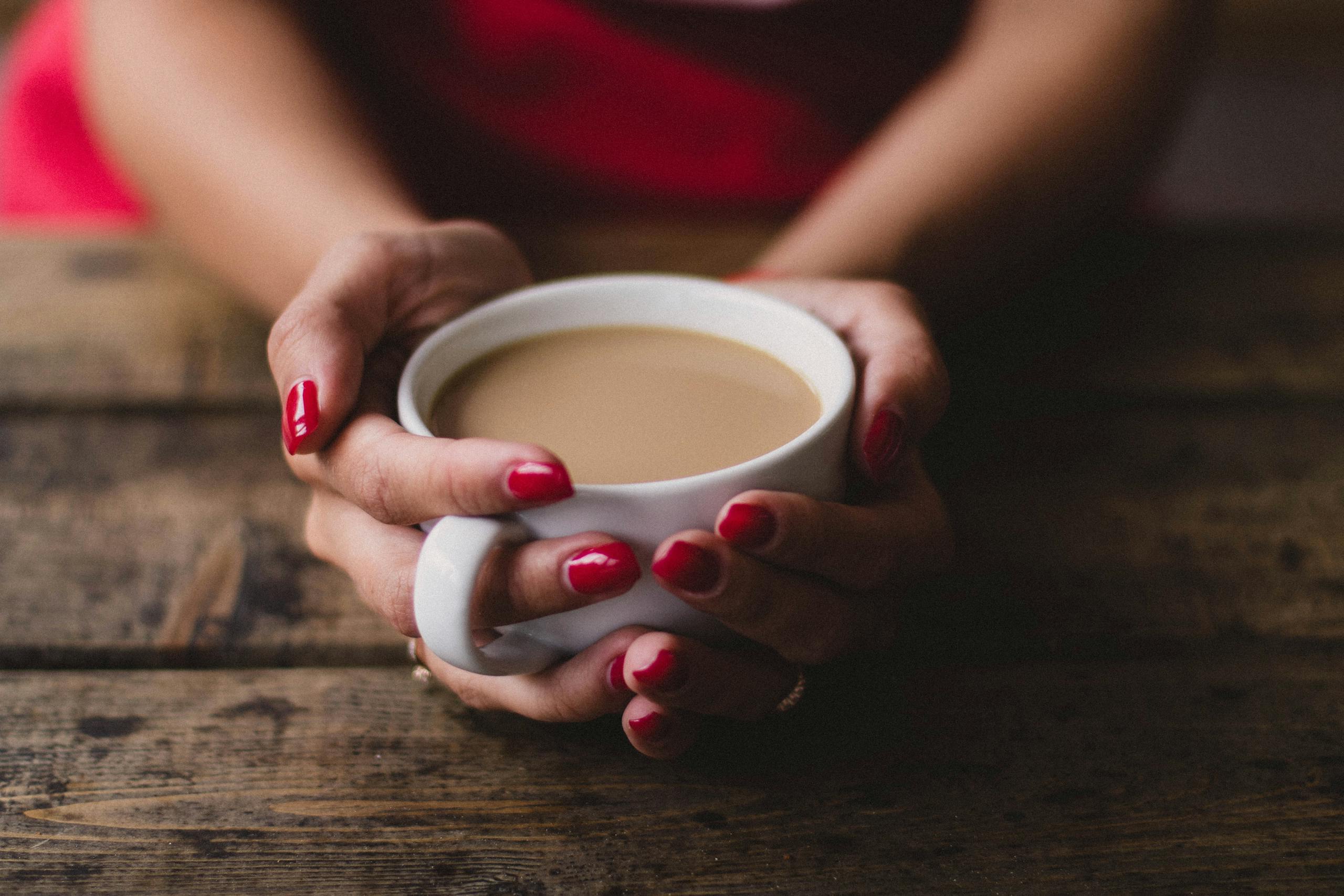 Warm indoor scene of a woman holding a steaming cup of coffee with red nails.